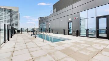 Uma área de piscina no terraço com vista desimpedida para o horizonte da cidade, um edifício moderno com grandes janelas e uma piscina com guarda-corpos de segurança.
