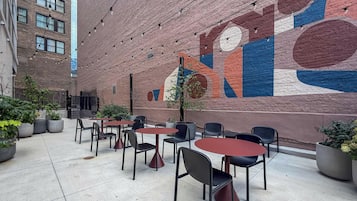 Área externa com mesas e cadeiras, parede de tijolos com mural e plantas em vasos.