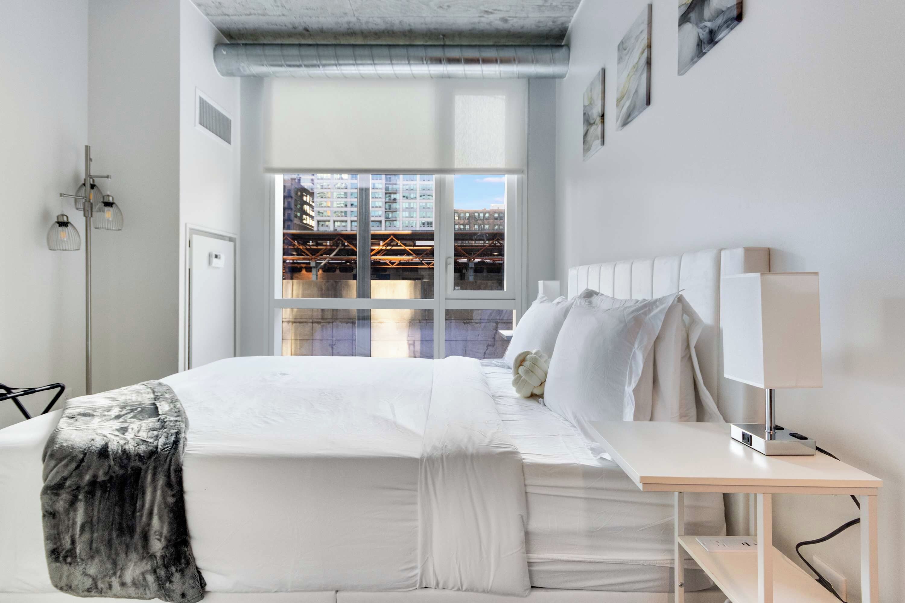 Un dormitorio moderno con una cama grande, una mesita de noche, una lámpara y vistas a la ciudad a través de una ventana.