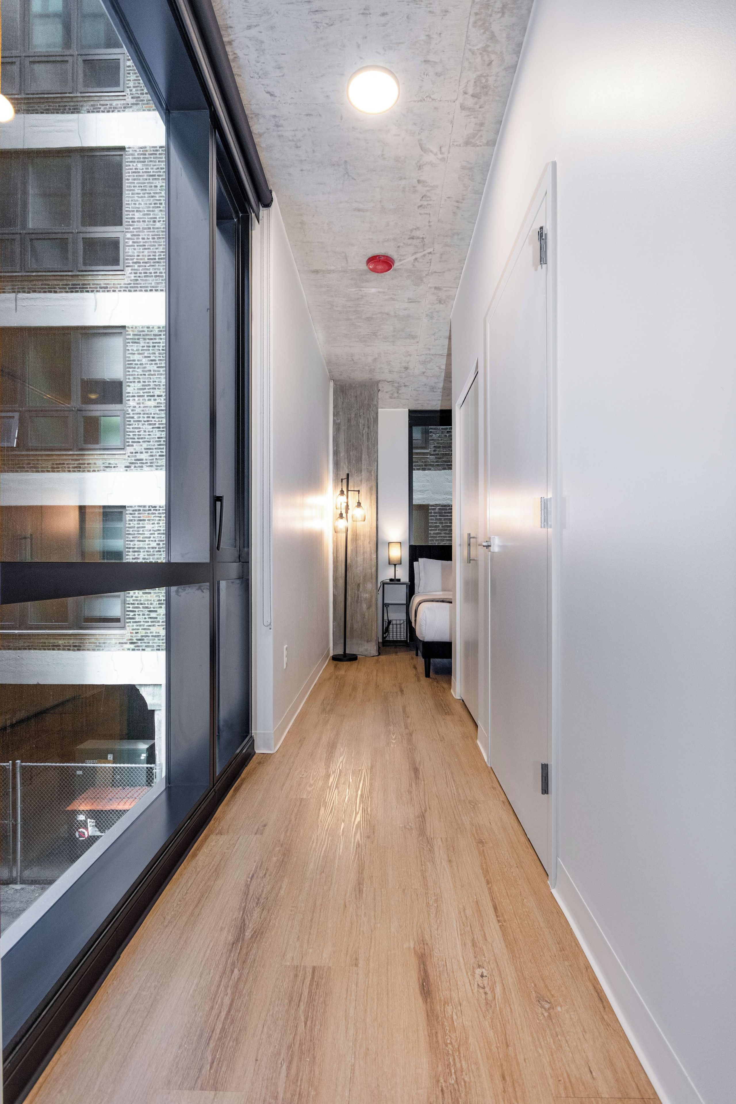 Um corredor estreito com piso de madeira, uma porta de vidro à esquerda e uma cama com mesa de cabeceira ao fundo.