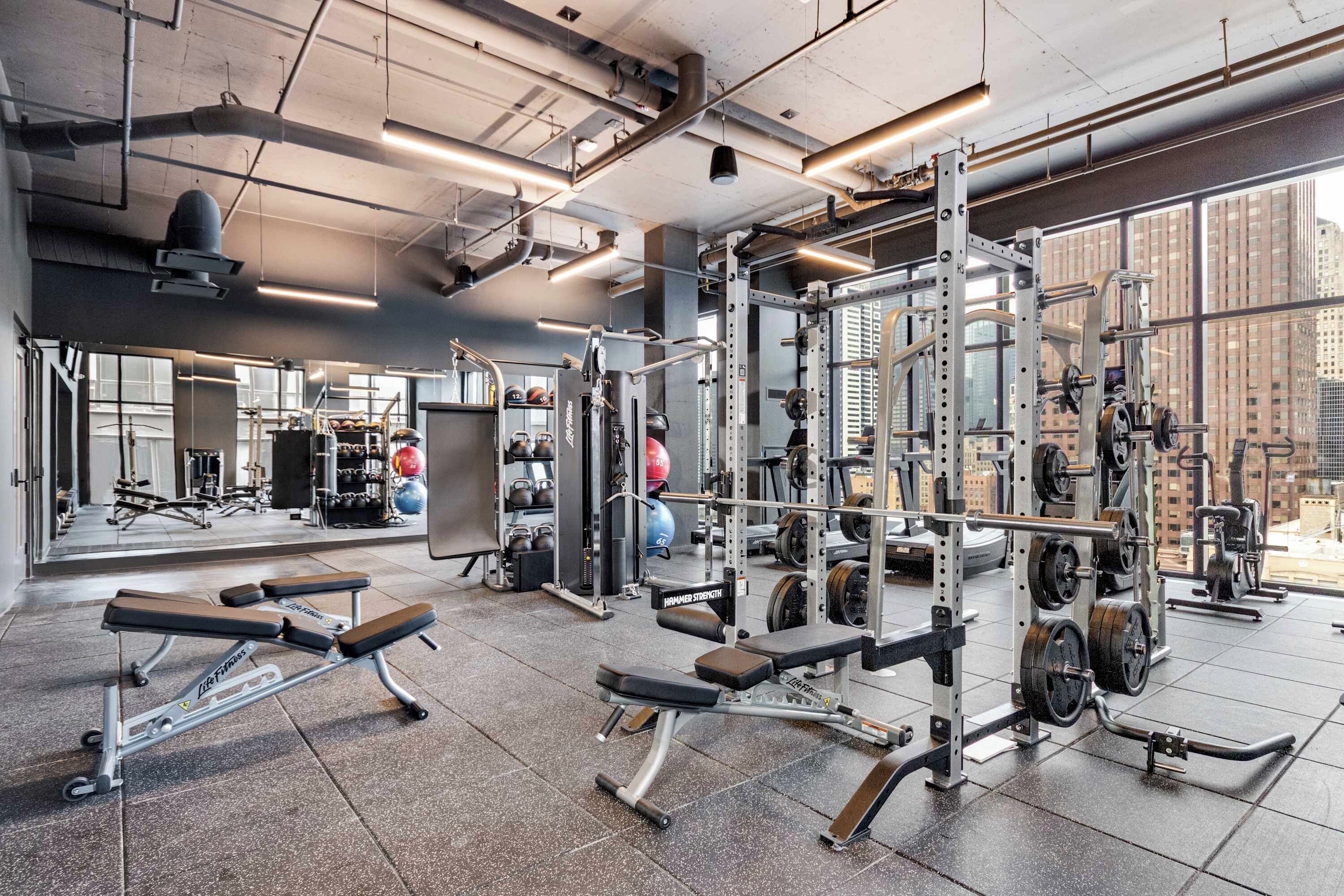 Uma academia com equipamentos de musculação, bancos e um espelho grande na parede.