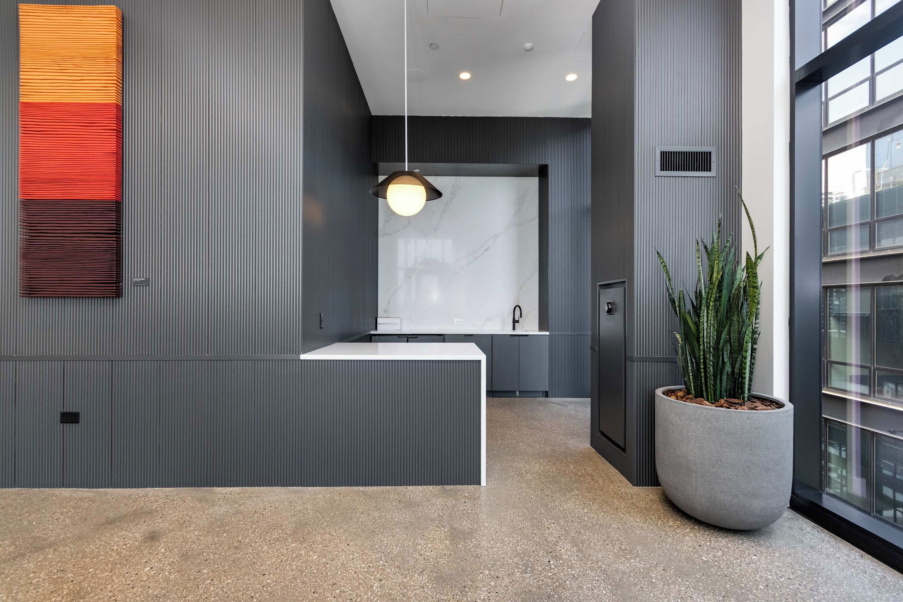 Uma área de recepção moderna, com um balcão branco, uma luminária pendente e uma grande planta em vaso.