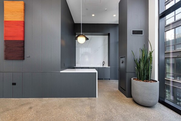 Uma área de recepção moderna, com um balcão branco, uma luminária pendente e uma grande planta em vaso.