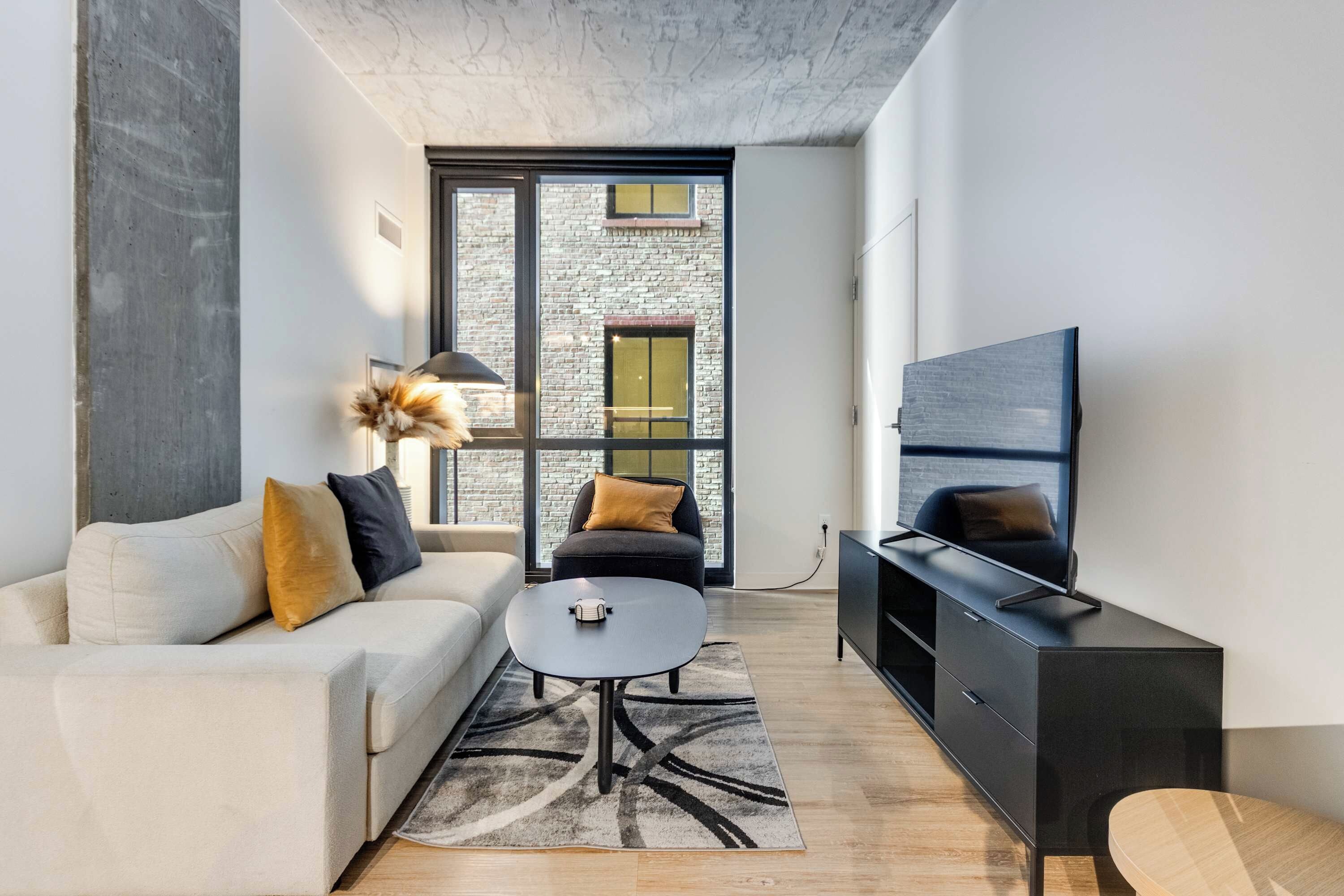 Sala de estar moderna com sofá, mesa de centro, TV e vista para um edifício de tijolos através de grandes janelas.