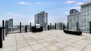 Terraço no telhado com churrasqueira, piso ladrilhado e vista dos edifícios altos ao redor.