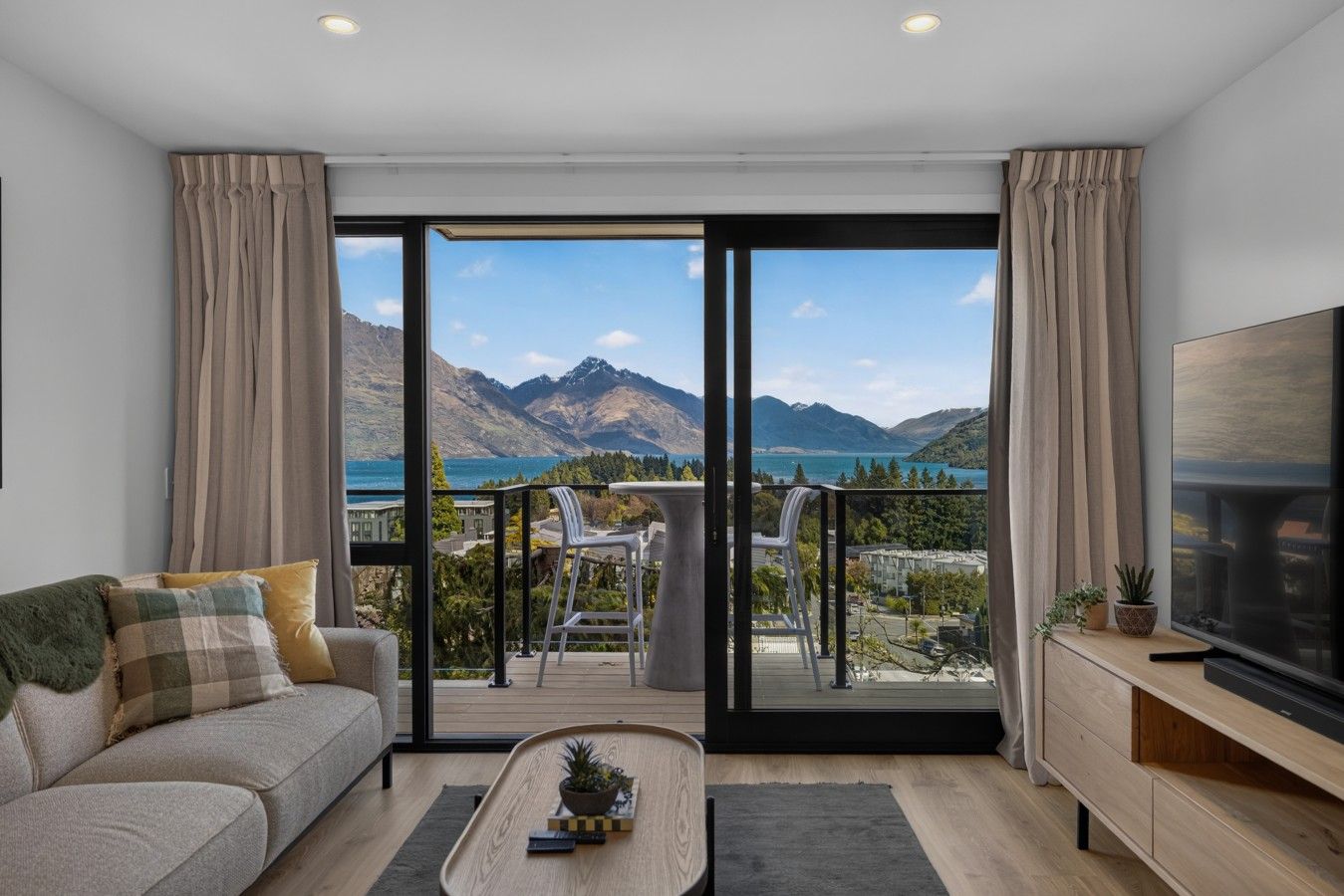 A modern living room with a sofa, a coffee table, and a TV on a wooden stand. The room has large sliding glass doors leading to an outdoor balcony with a view of mountains and a lake.