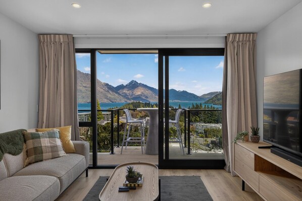 Una sala de estar moderna con un sofá, una mesa de centro y un televisor sobre un mueble de madera. La habitación tiene grandes puertas correderas de cristal que dan a un balcón exterior con vistas a montañas y un lago.
