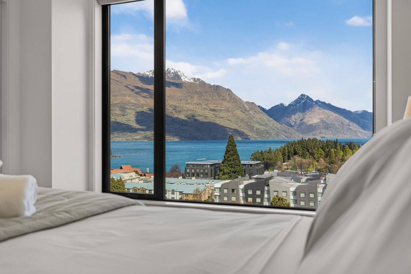 A hotel room with a large window offering a view of a lake, mountains, and a cityscape.