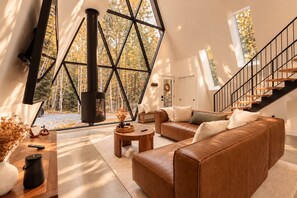 A modern interior with a large window, a fireplace, a sofa, and a staircase. - Cozy Peak | Luxury A-Frame | Hot Tub | Lake Access (Chertsey)