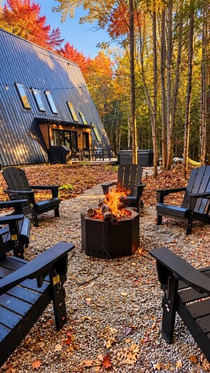 A cabin with a fire pit surrounded by Adirondack chairs, set in a forest with autumn leaves. - Cozy Peak | Luxury A-Frame | Hot Tub | Lake Access (Chertsey)