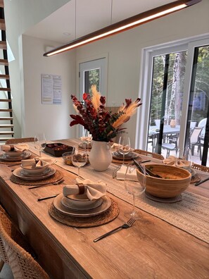 Una mesa de comedor dispuesta con platos, cuencos y cubiertos, un florero con flores y vistas del exterior a través de amplias ventanas.