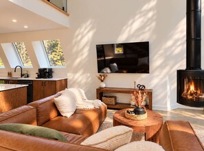 A modern living room with a leather sofa, a flat-screen TV, a fireplace, and a kitchen area with a sink and coffee machine. - Cozy Peak | Luxury A-Frame | Hot Tub | Lake Access (Chertsey)