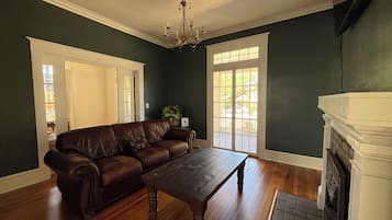 A room with a brown leather sofa, a wooden coffee table, a white fireplace, and a large window with a view of the outdoors.