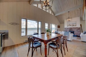 A spacious living area with a large wooden dining table, comfortable seating, a fireplace, and large windows offering a view of the water. - Le Tomahawk - Potton en Estrie (Potton)