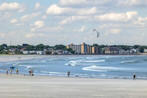 Una playa con gente, una cometa y edificios al fondo.