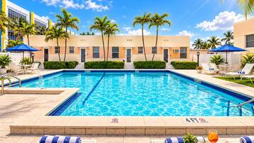 Una piscina con sillones de descanso y sombrillas, palmeras y un edificio al fondo.