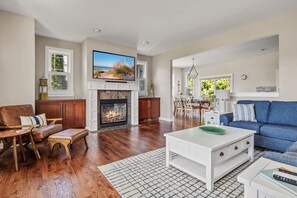A living room with a fireplace, a television mounted above it, a blue sofa, and a dining area in the background. - Keppel Cove | Saugatuck • Hot Tub • Rec Room! (Saugatuck)