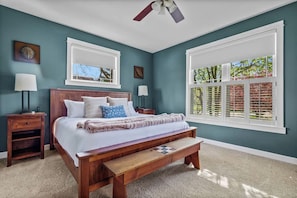 A bedroom with a wooden bed, two bedside tables, a bench, a ceiling fan, and a large window with shutters. - Keppel Cove | Saugatuck • Hot Tub • Rec Room! (Saugatuck)