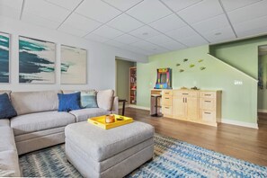 Een woonkamer met een bank, voetenbankje en salontafel. Er is een boekenkast en een kleine barruimte met een spiegel.