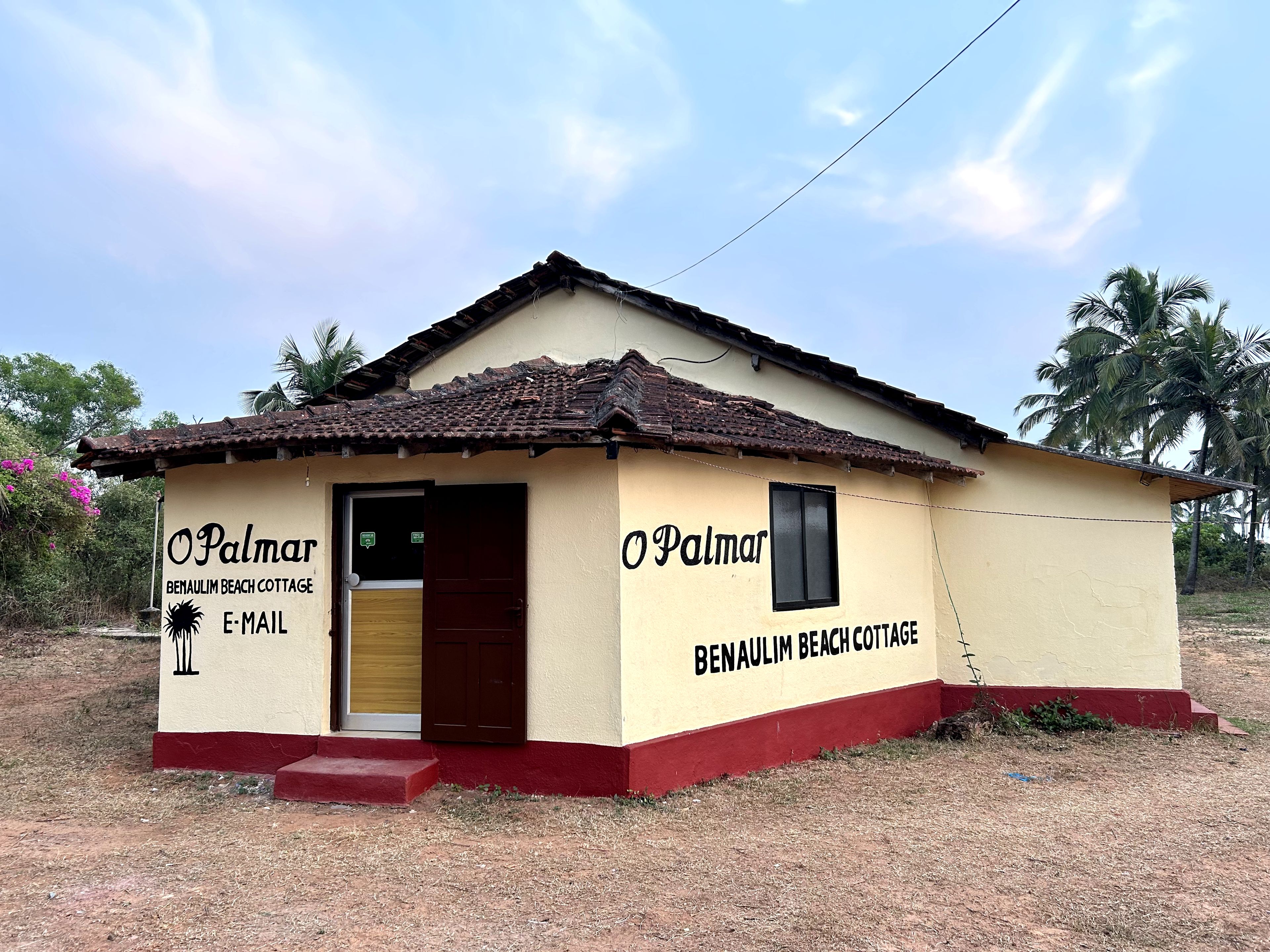A small, single-story building with a tiled roof and a red base, labeled 'Benaulim Beach Cottage.'