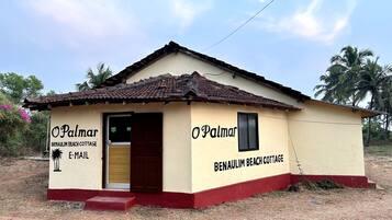 A small, single-story building with a tiled roof and a red base, labeled 'Benaulim Beach Cottage.'