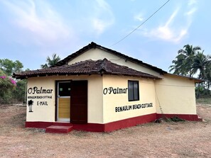Um pequeno edifício térreo com telhado telhado e base vermelha, com a placa 'Benaulim Beach Cottage'.