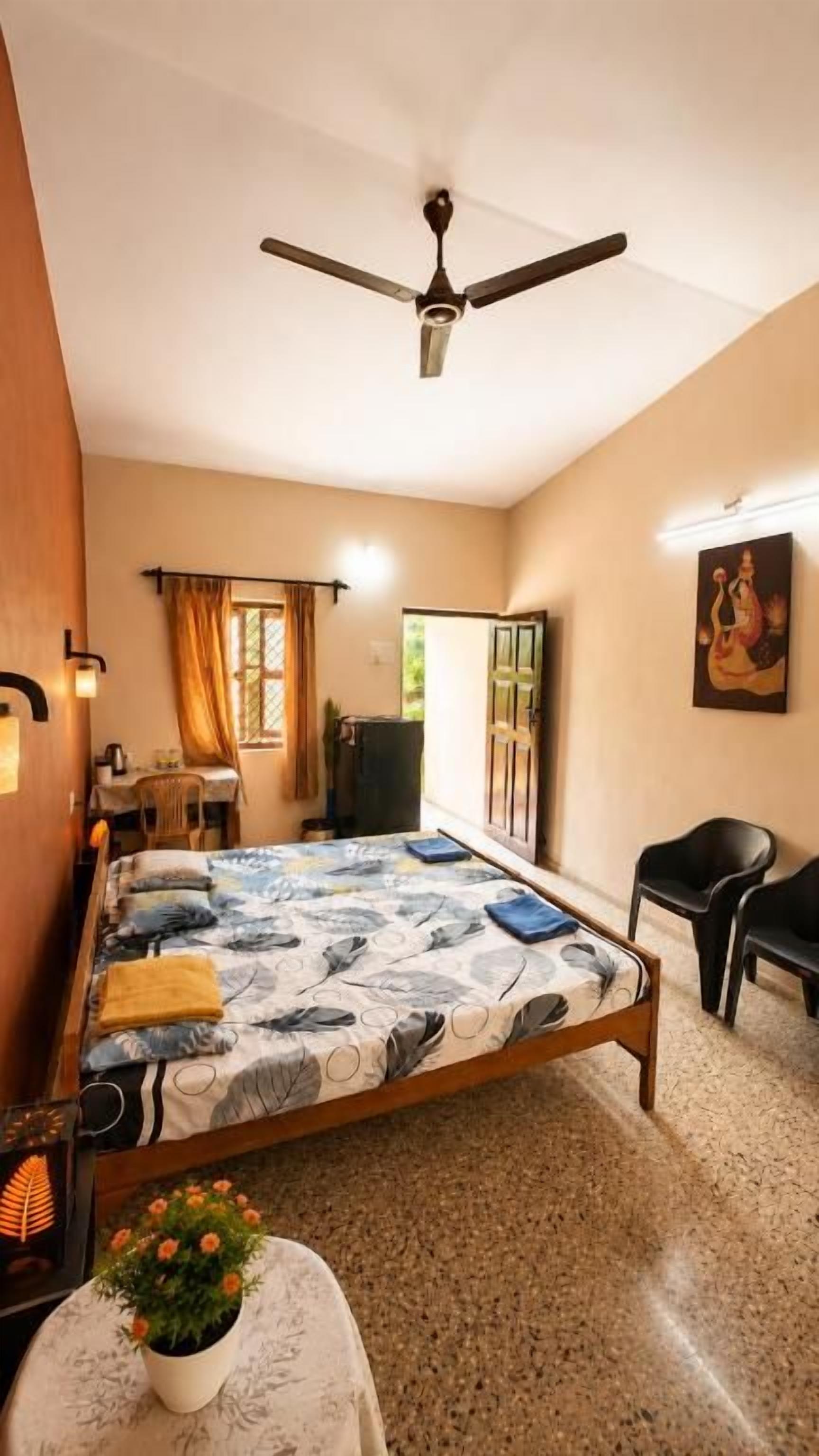 A bedroom with a bed, a ceiling fan, a desk with a chair, a potted plant, and a painting on the wall.