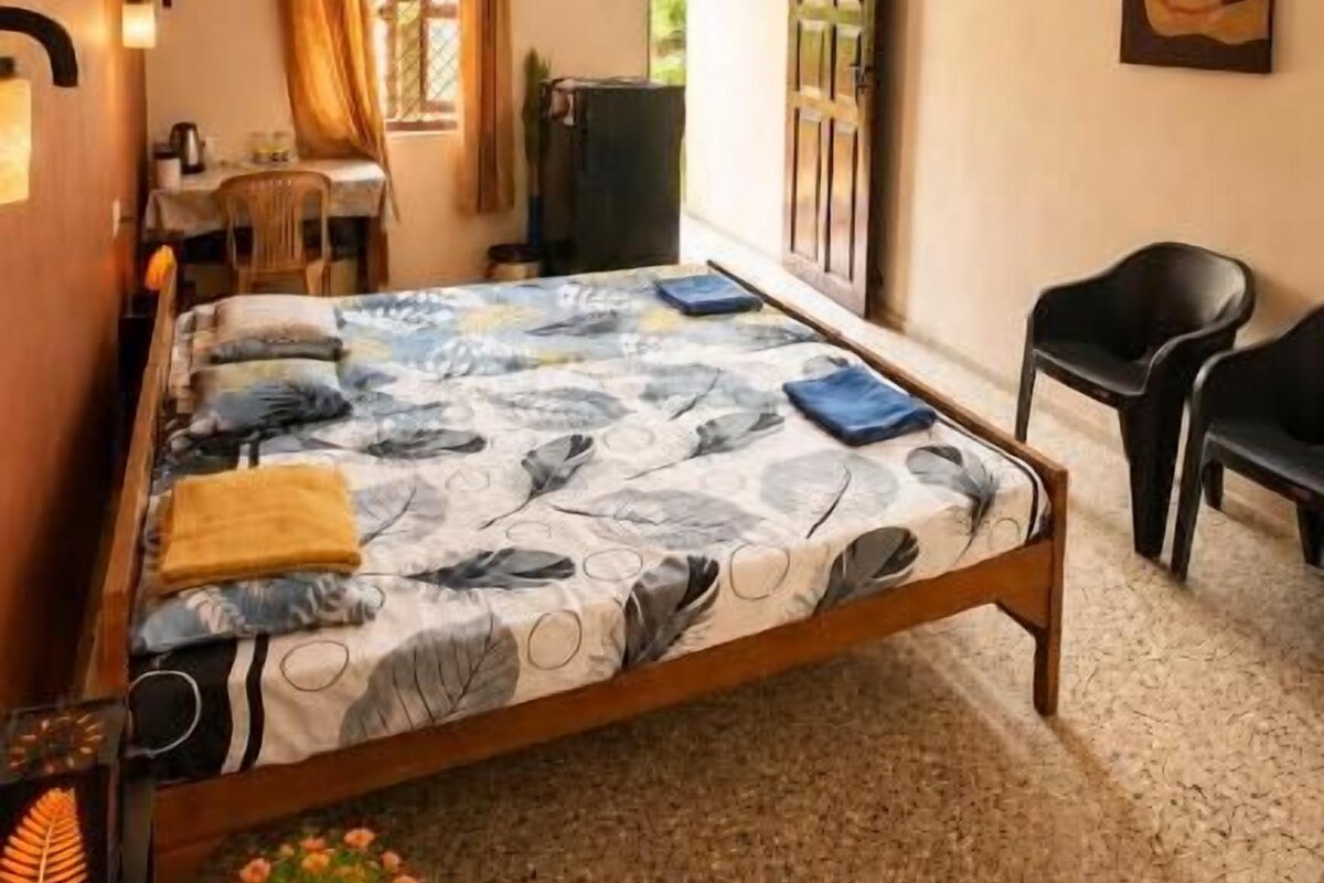 Quarto com cama, ventilador de teto, escrivaninha com cadeira, planta em vaso e um quadro na parede.