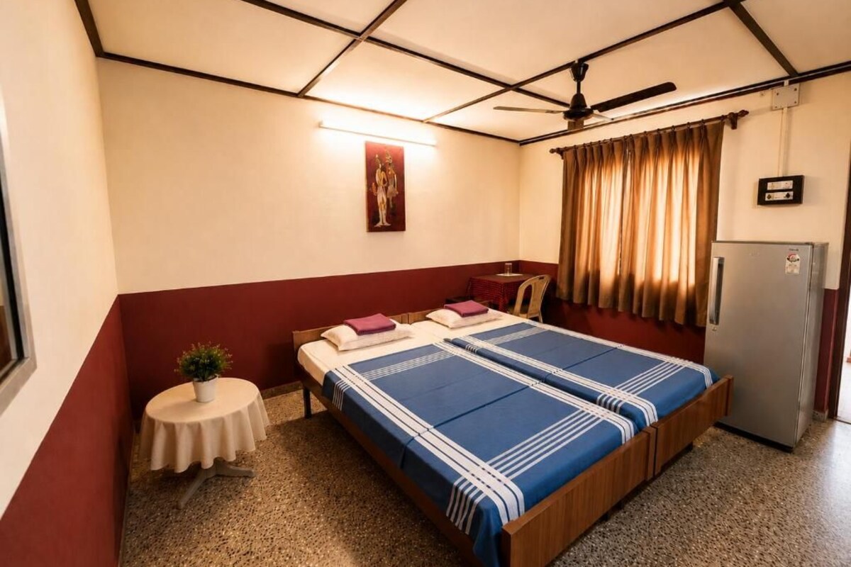 Quarto individual com uma cama de madeira, uma mesinha com uma planta, um ventilador de teto e uma geladeira.