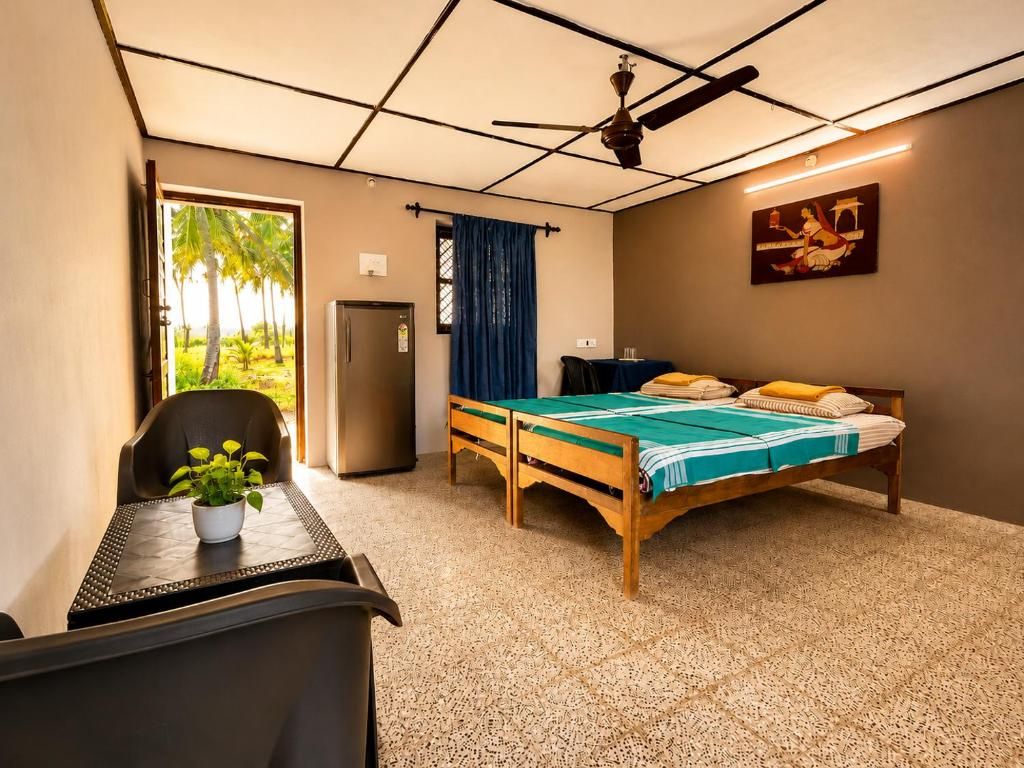 Una habitación con cama, escritorio, silla, planta en maceta, ventilador de techo y ventana con vista a palmeras.