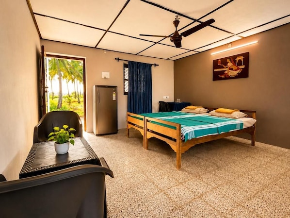 Una habitación con cama, escritorio, silla, planta en maceta, ventilador de techo y ventana con vista a palmeras.