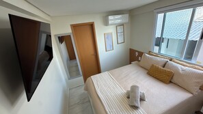 A hotel room with a bed, a TV mounted on the wall, a window with a view of the roof, and an air conditioning unit. - TNP - Vila Porto 310 (Ipojuca)