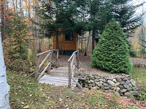 A wooden cabin with a wooden bridge leading to it, surrounded by trees and a stone wall.