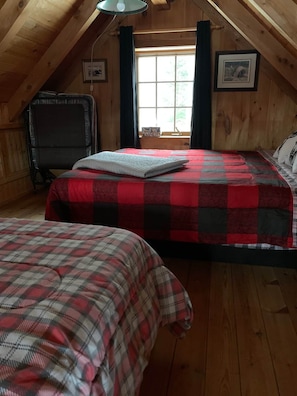 A cozy attic bedroom with two beds, wooden walls, a window with curtains, and a framed picture on the wall.