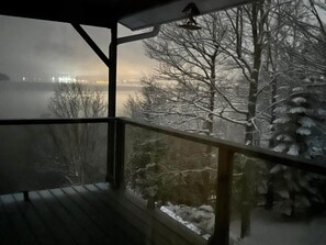 A view from a covered balcony with a railing, overlooking a snowy landscape and a body of water with distant lights. - Maison du Lac (Témiscouata-sur-le-Lac)
