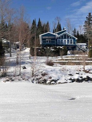 A two-story house with a blue exterior, surrounded by snow-covered ground and trees. - Maison du Lac (Témiscouata-sur-le-Lac)