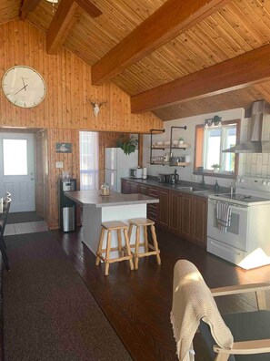 A wooden interior with a kitchen area, including a refrigerator, stove, and wooden stools. - Maison du Lac (Témiscouata-sur-le-Lac)