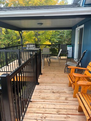 A wooden deck with a grill, chairs, and a covered area. - Maison du Lac (Témiscouata-sur-le-Lac)
