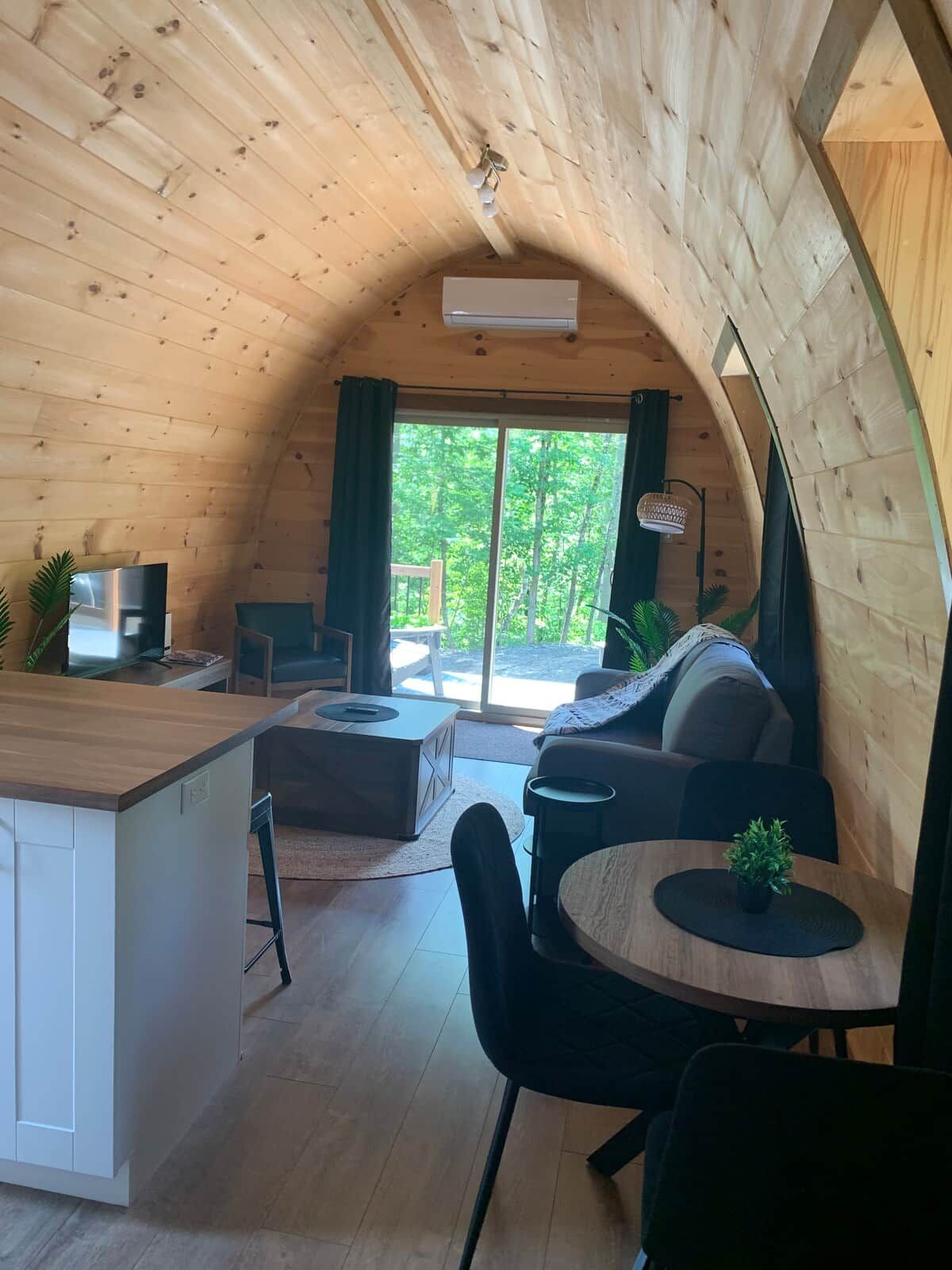 A cozy cabin interior with a wooden ceiling, a dining area, a sofa, and a kitchen area.