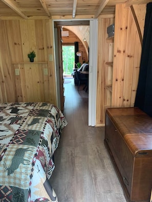 A wooden interior with a bed, a bench, and a desk area with a chair and a lamp.