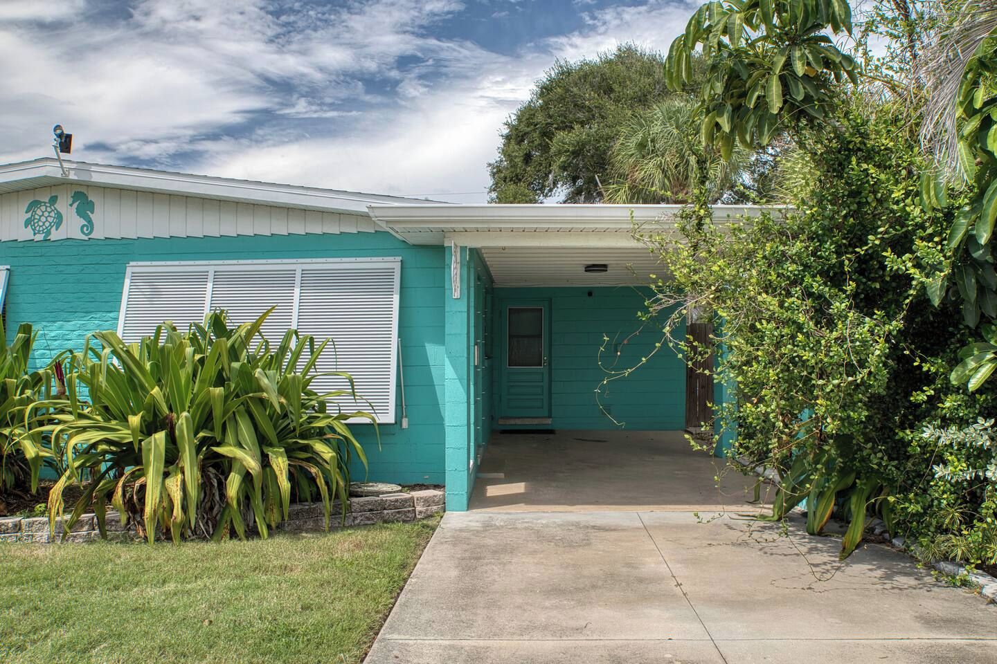 Une maison de plain-pied à la façade de couleur turquoise, dotée d’un porche et d’un jardin devant, où l’on trouve des plantes tropicales.