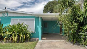 Une maison de plain-pied à la façade de couleur turquoise, dotée d’un porche et d’un jardin devant, où l’on trouve des plantes tropicales.