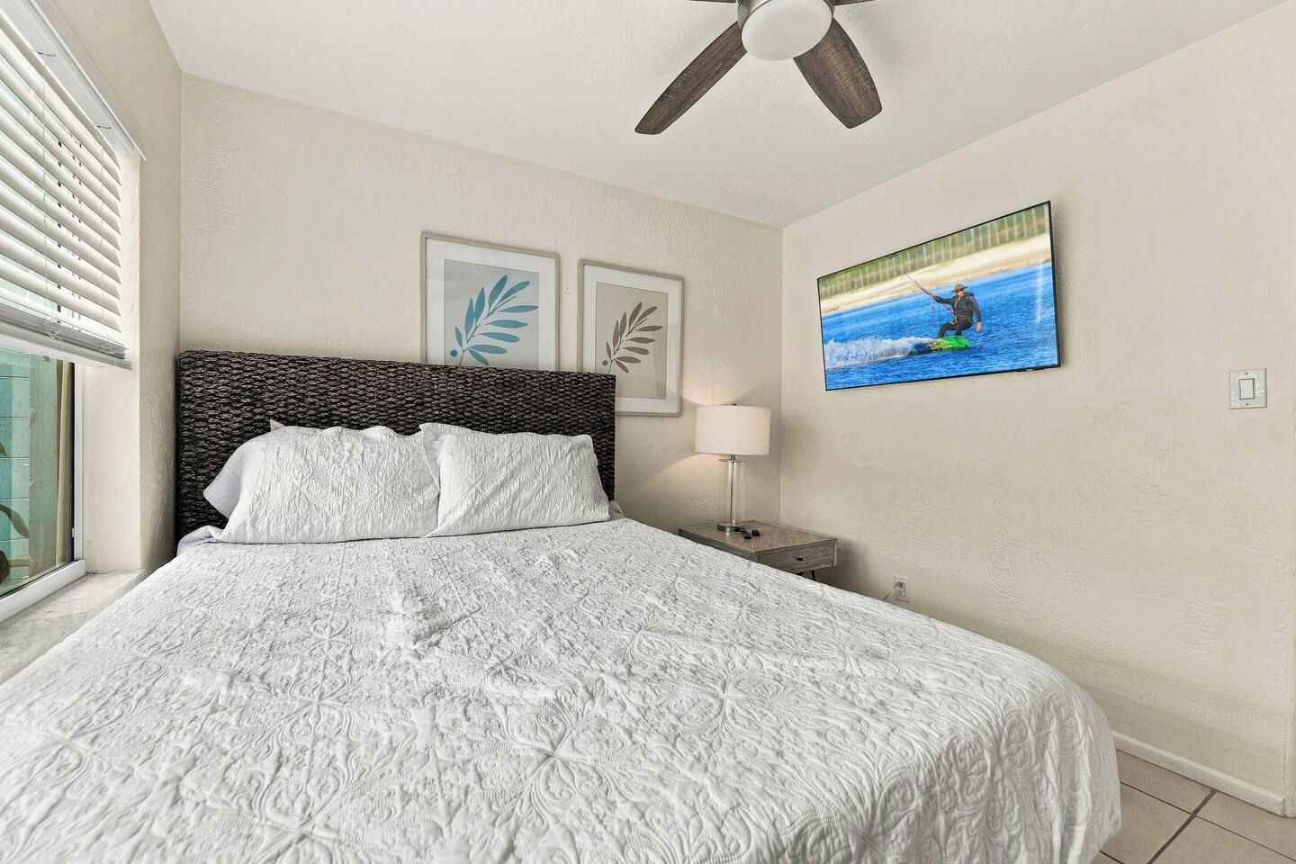 A bedroom with a large bed, a ceiling fan, a nightstand with a lamp, and a framed picture on the wall.