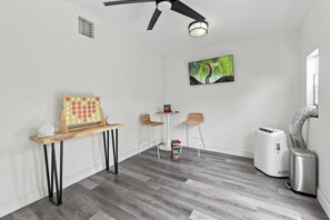 Une pièce dotée d’un ventilateur de plafond, d’un téléviseur fixé au mur, d’une table en bois sur laquelle se trouve un puzzle, de deux tabourets de bar et d’un purificateur d’air blanc.