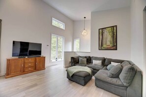 A spacious living room with a large flat-screen TV, a wooden entertainment unit, a grey sectional sofa, and a painting of a forest. - La Catherine  - Weedon (Weedon)