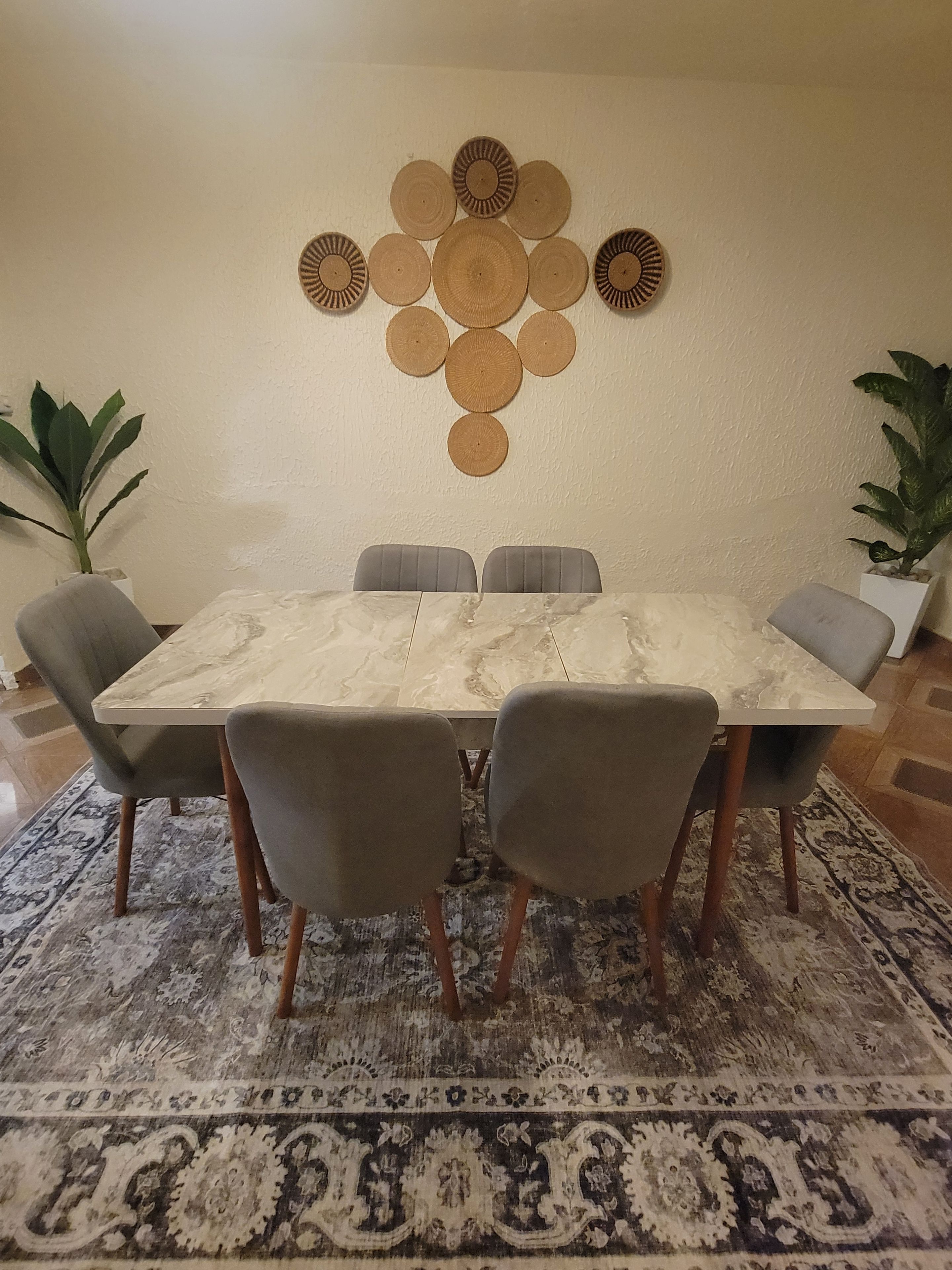 A dining area with a marble table, four grey upholstered chairs, a decorative wall hanging, and a patterned rug.