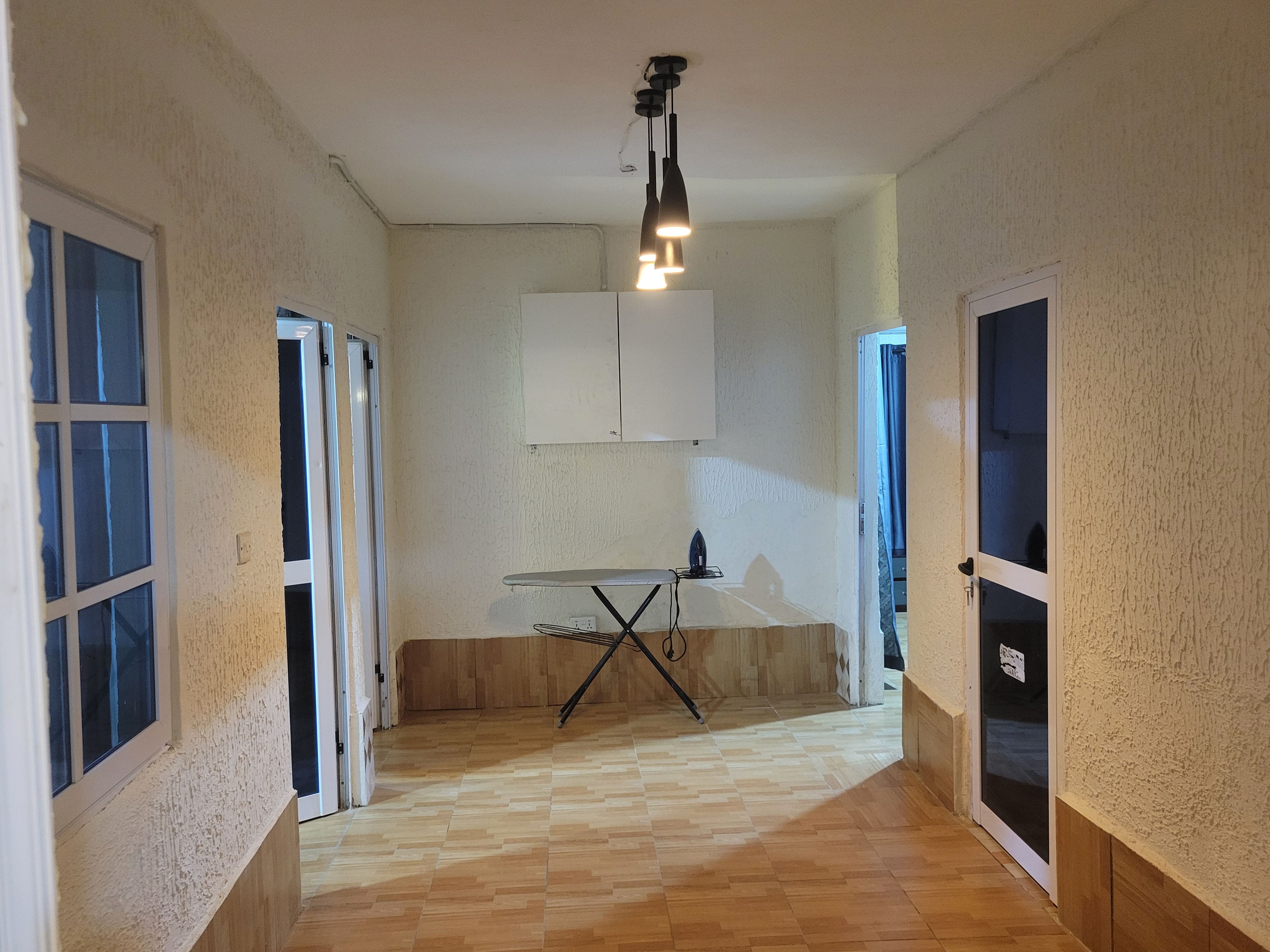 A hallway with a wooden floor, a table with an iron, and a ceiling light fixture.