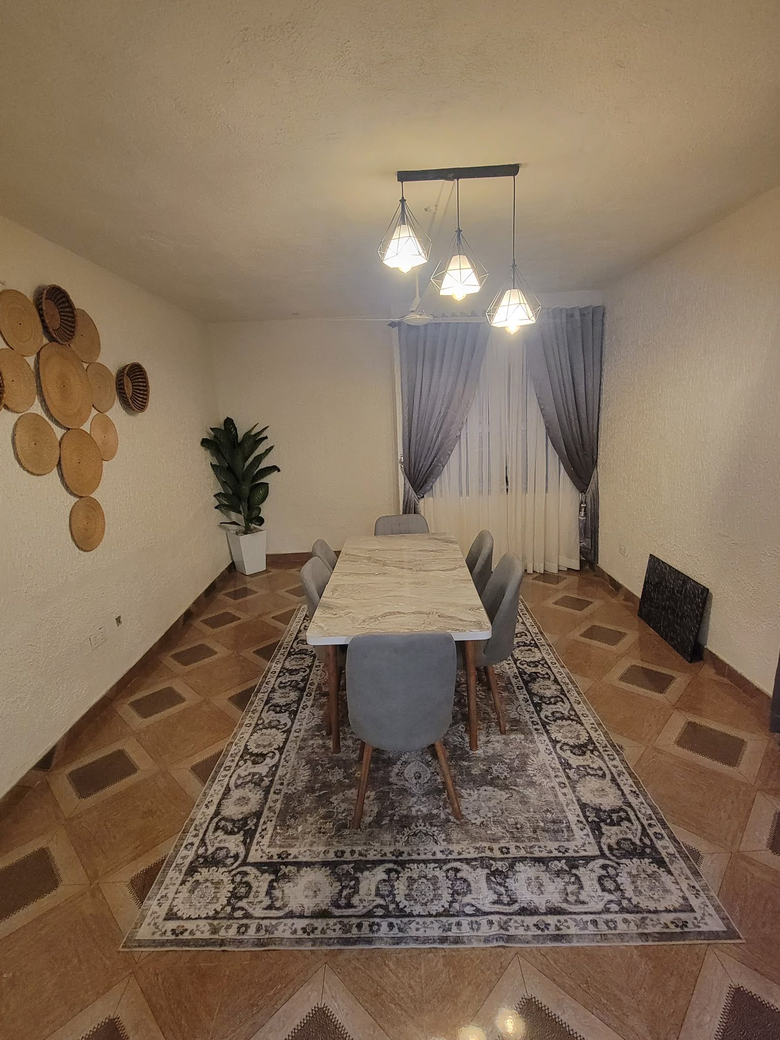 A dining area with a long table, chairs, a patterned rug, hanging lights, a potted plant, and wall decorations.