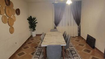 A dining area with a long table, chairs, a patterned rug, hanging lights, a potted plant, and wall decorations.
