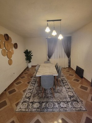 Un comedor con una mesa larga, sillas, alfombra estampada, lámparas colgantes, una planta en maceta y adornos en la pared.